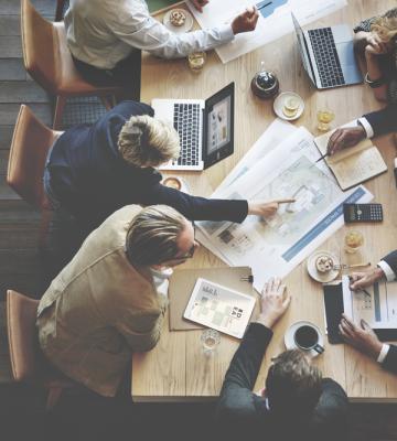 Professionals at a large table with laptops, coffees, and meeting notes.