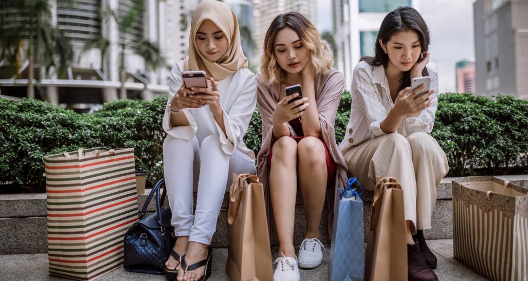 three girls sit on their phones with shopping bags after a long day of shopping