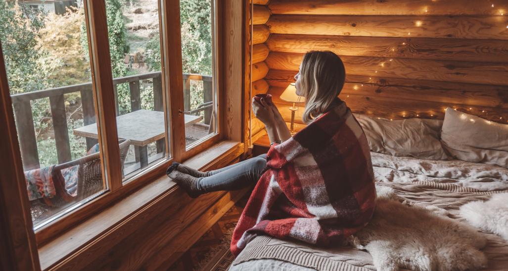 Woman in cosy log cabin looking outside holding mug
