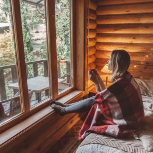 Woman in cosy log cabin looking outside holding mug
